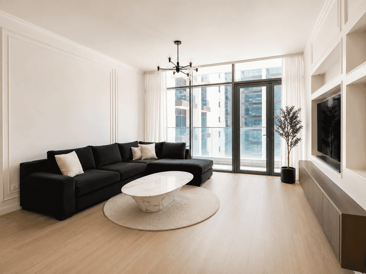 Dubai Hills renovation showing a completed apartment living room with wall moulding, black L-shaped sofa, marble oval coffee table, custom TV shelving wall and floor-to-ceiling balcony windows by Fitout Renovation Dubai