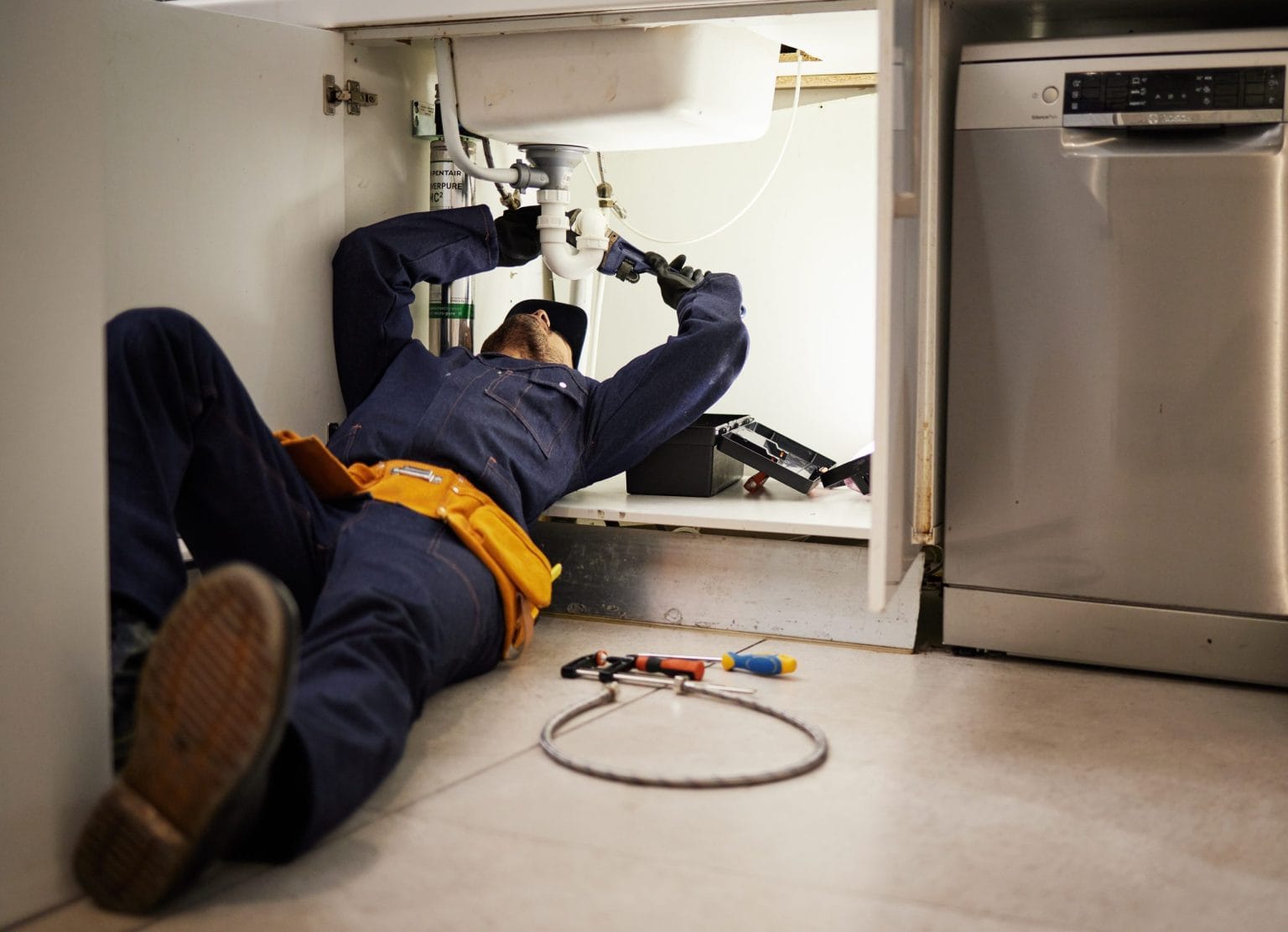 Plumber working under a kitchen sink beside a dishwasher in Dubai