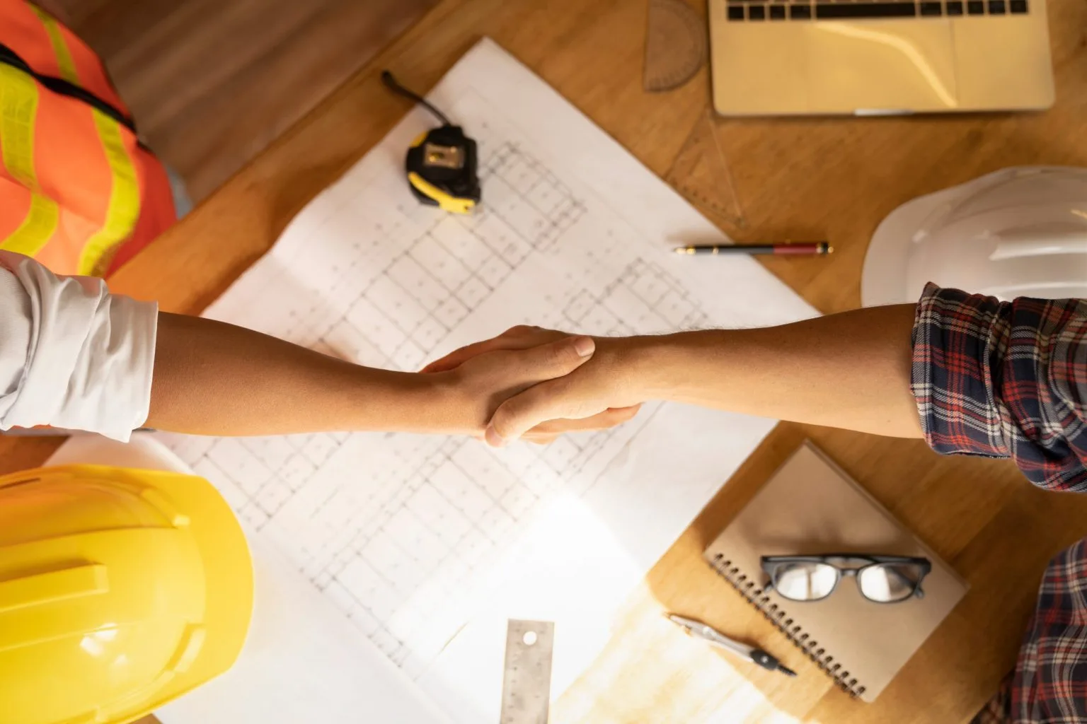 Client and project lead shaking hands over drawings, a tape measure and a hard hat